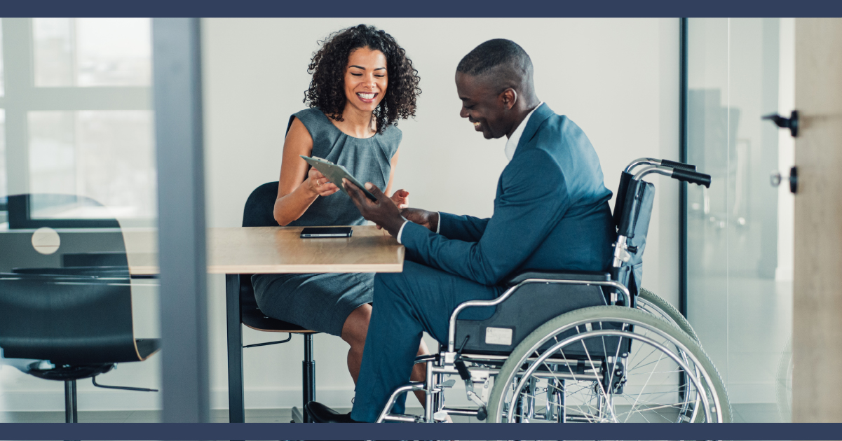 Man in suit in wheelchair at desk, signing paperwork presented by a woman.
