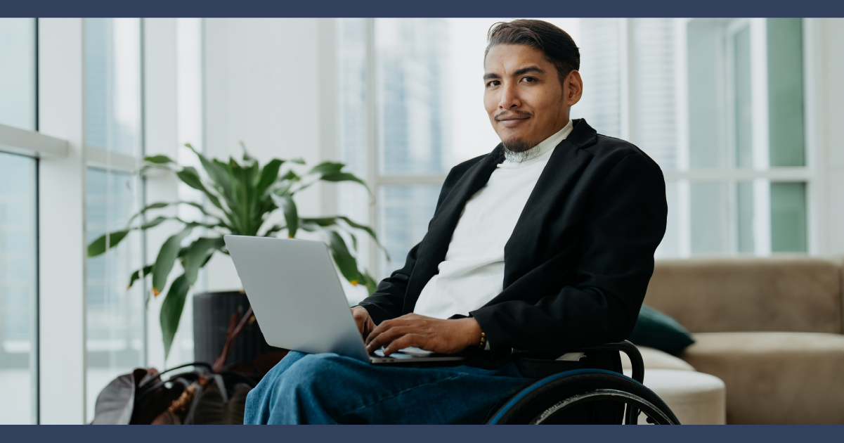 man wearing shirt and suit jacket, sitting in a wheelchair in an office, typing on a alptop computer