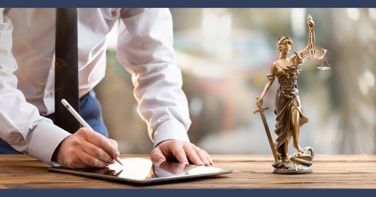 Man leaning over a desk signing legal paperwork with a 'lady justice' model statue on the desk nearby
