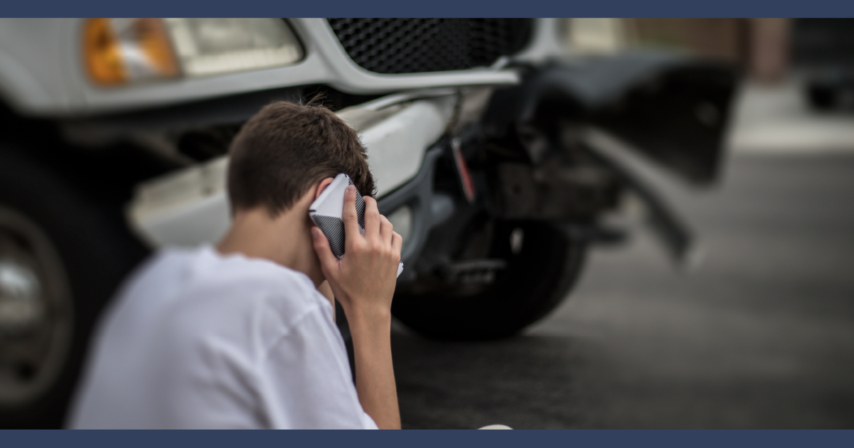 View of sitting man using his cell phone to make a call after a car accident, in the background is a car with damaged fender.