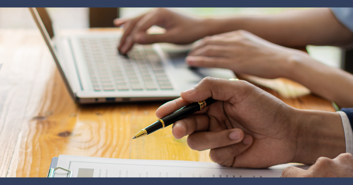 close up of hand holding a pen, with a desk and laptop in the background of a meeting