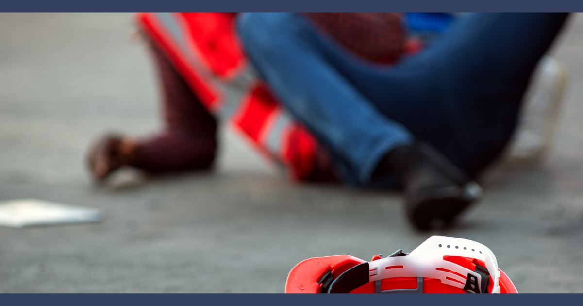 Person on the floor injured, wearing hi-visability jacket and their hardhat is on the floor as if they have fallen over.