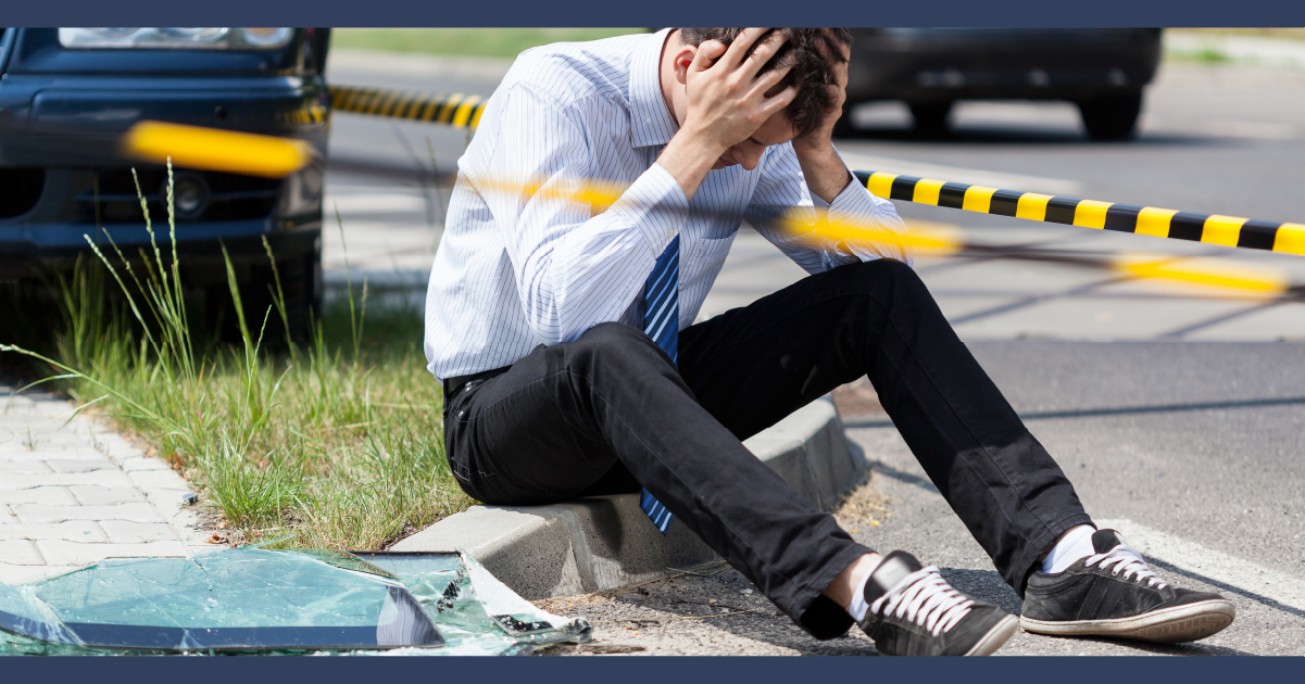Pale man an in suit with his head in his hands sitting on the kerb with car accident scene behind him