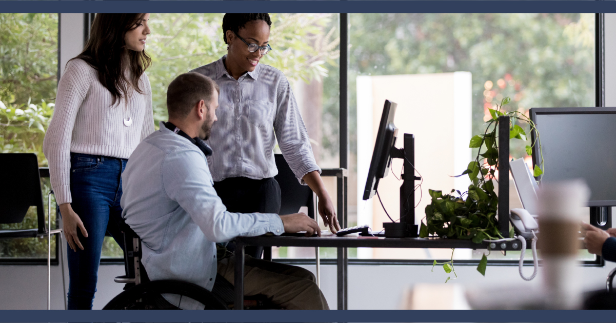 two office workers standing next to colleague who is in a wheelchair, all looking at a computer screen